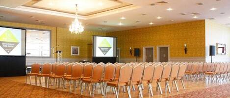 A conference room with rows of chairs, a carpeted floor, a chandelier, and a projector screen.