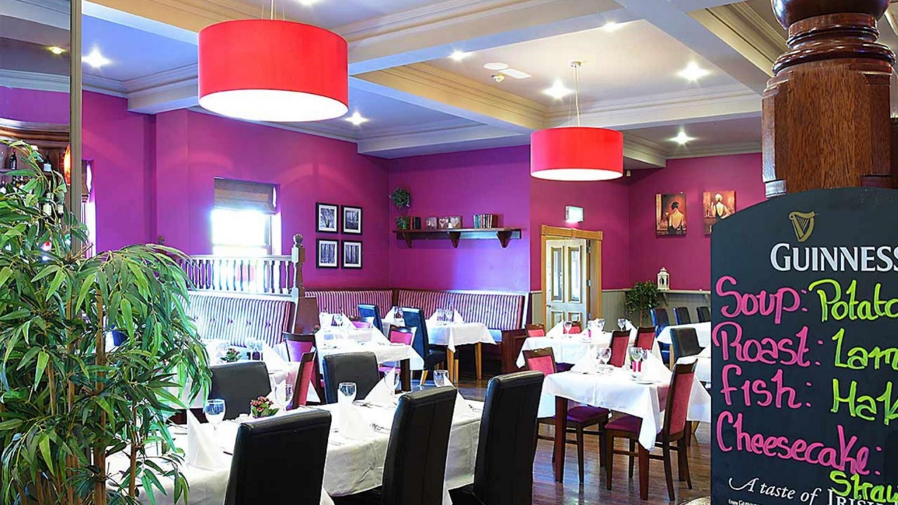 Un ristorante con pareti viola, luci a sospensione rosse e una lavagna con il menù che offre zuppa, arrosto, pesce e cheesecake.