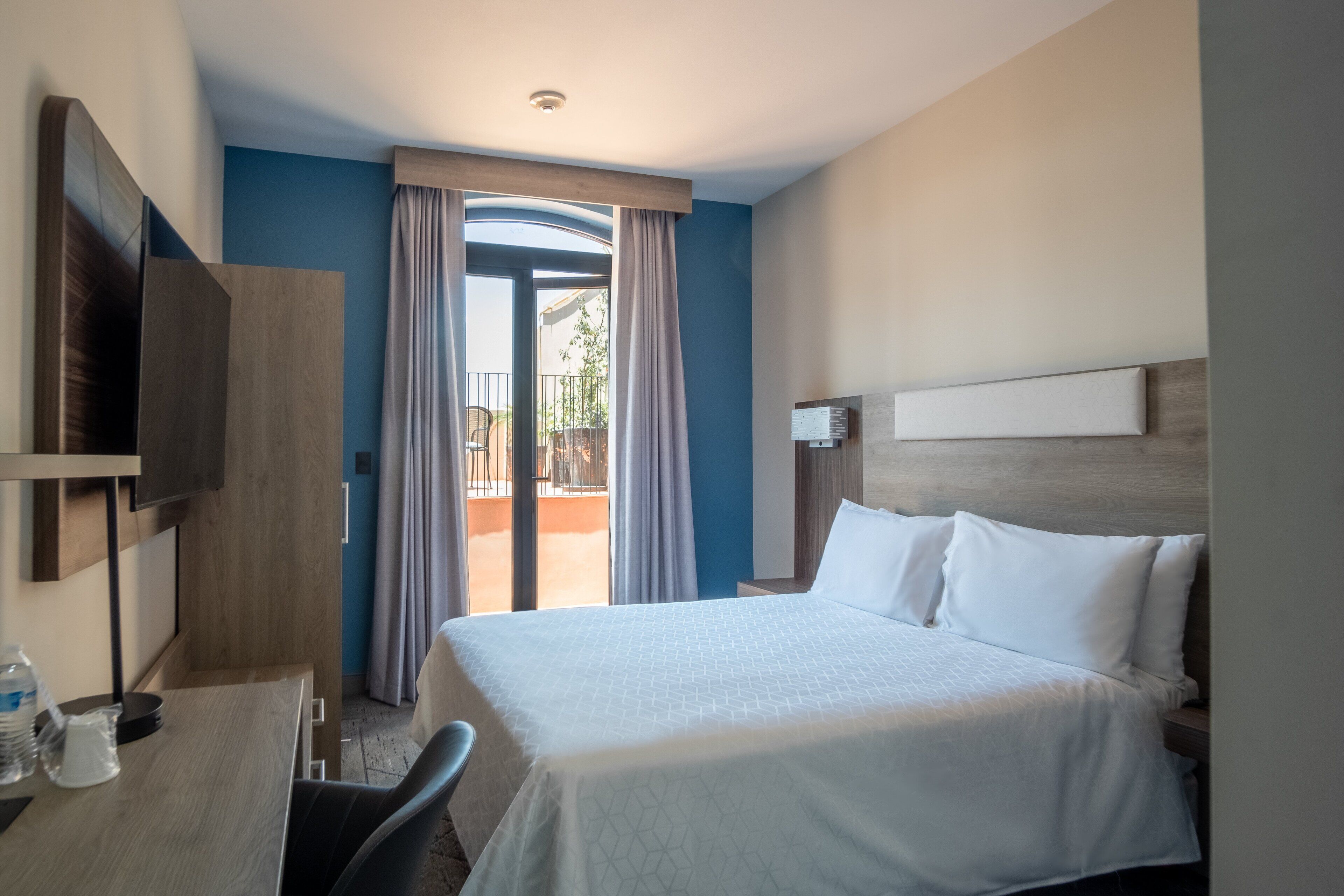 Quarto de hotel com uma cama, uma escrivaninha com cadeira, televisão e varanda com cortinas.
