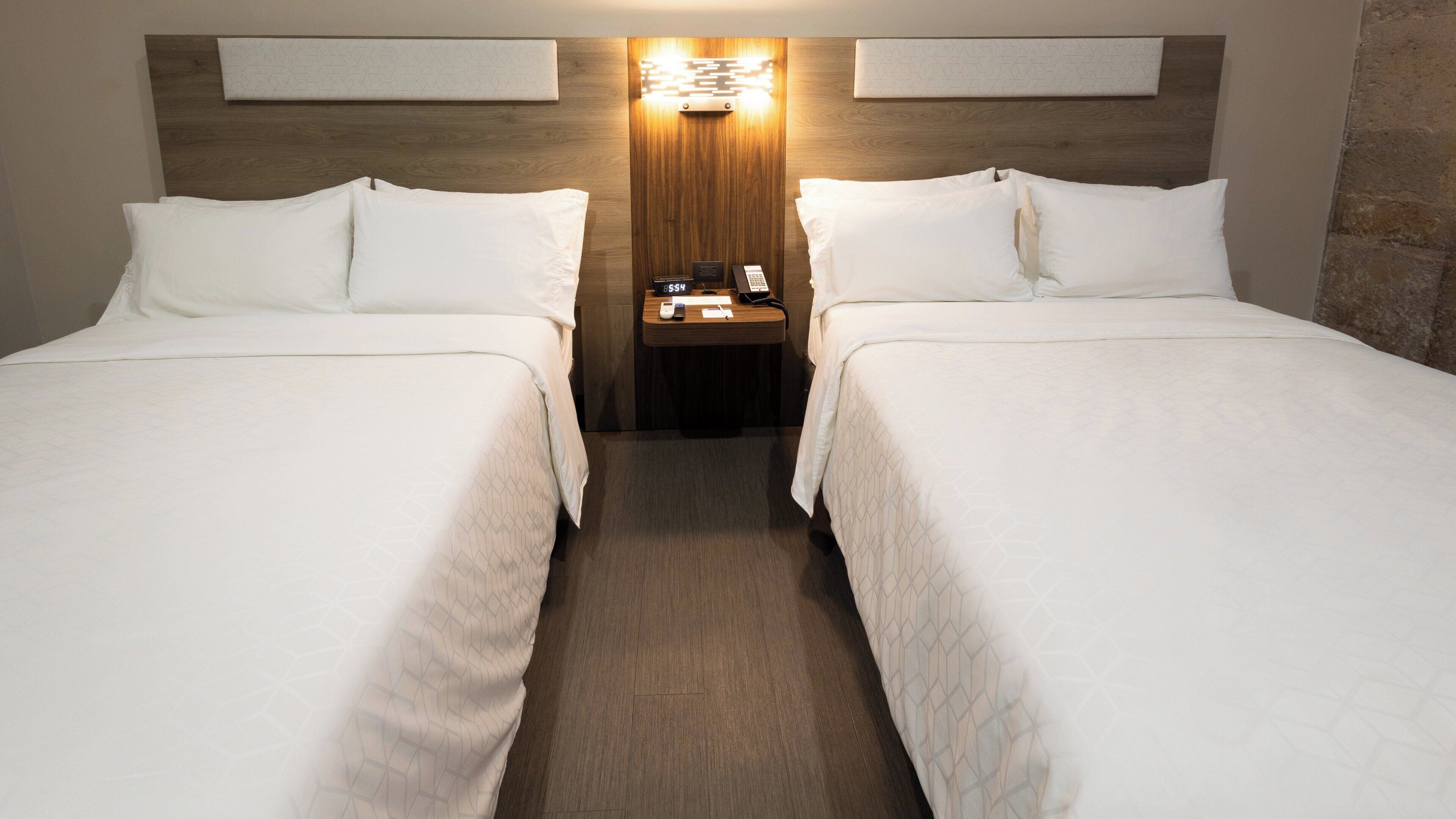 A hotel room with two beds, a nightstand with a phone and a lamp, and a wooden headboard with a geometric light fixture.