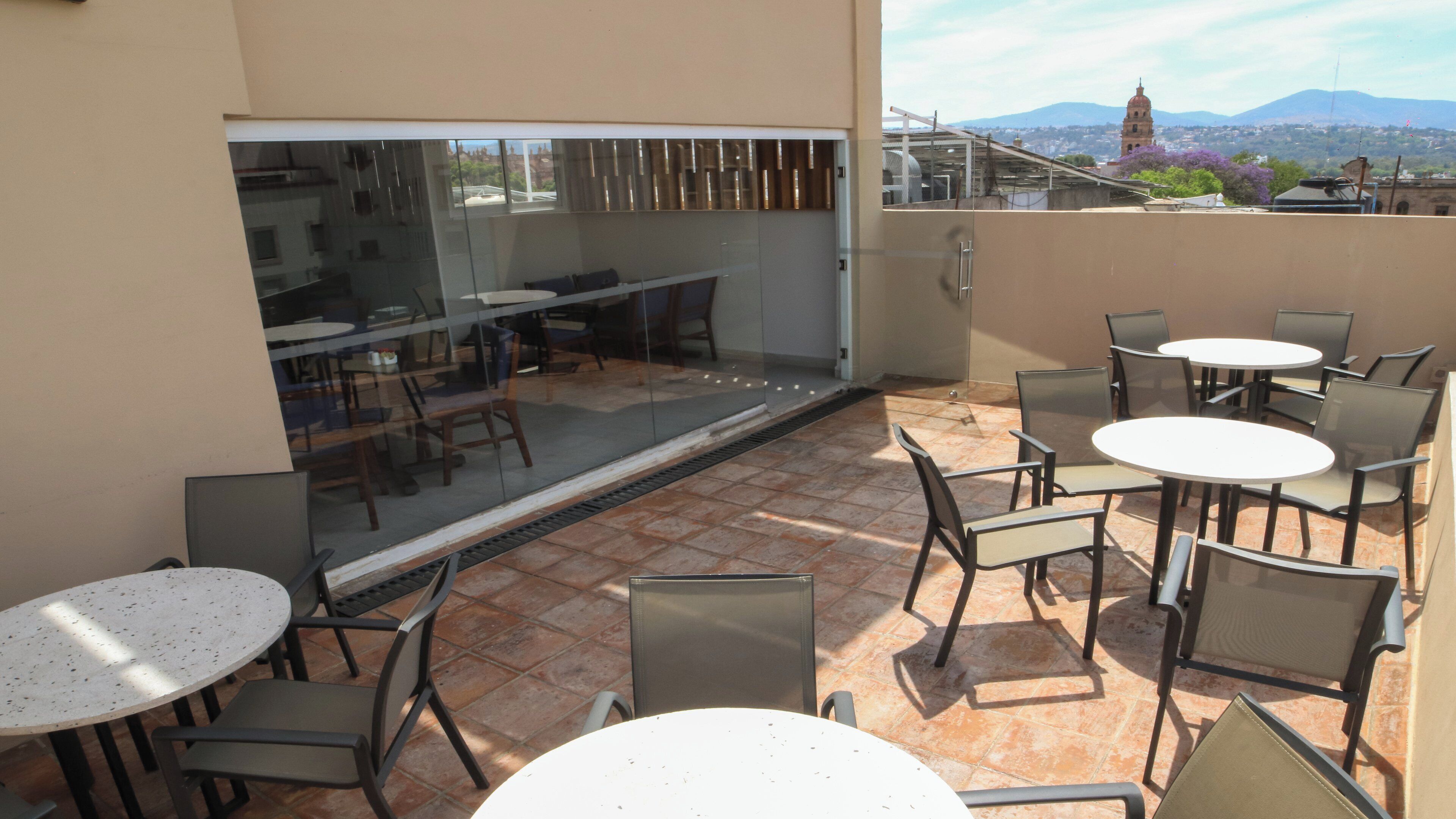 Un patio al aire libre con mesas y sillas redondas, un muro de cristal con vistas a un paisaje urbano con montañas al fondo.