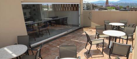 An outdoor patio with round tables and chairs, a glass wall overlooking a cityscape with mountains in the distance.