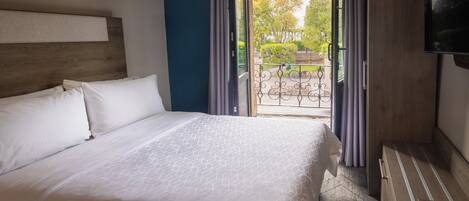 A hotel room with a large bed, a wooden headboard, a TV, and a balcony with a view of trees and a fence.