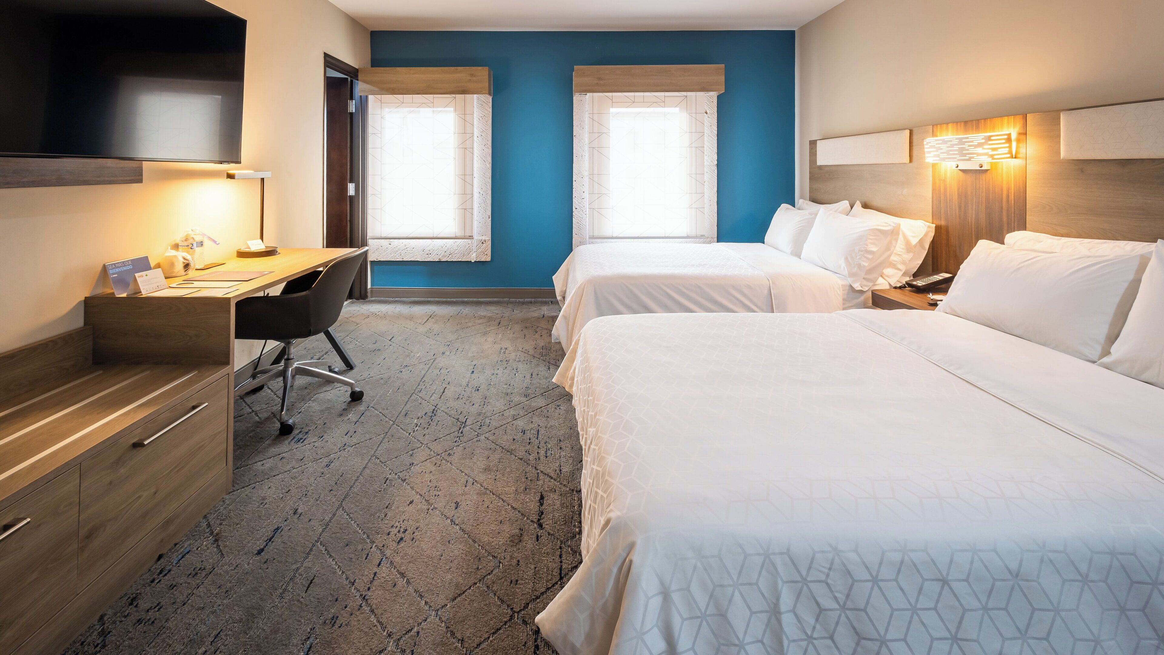 A hotel room with two beds, a desk, and a chair. There are two windows with curtains.