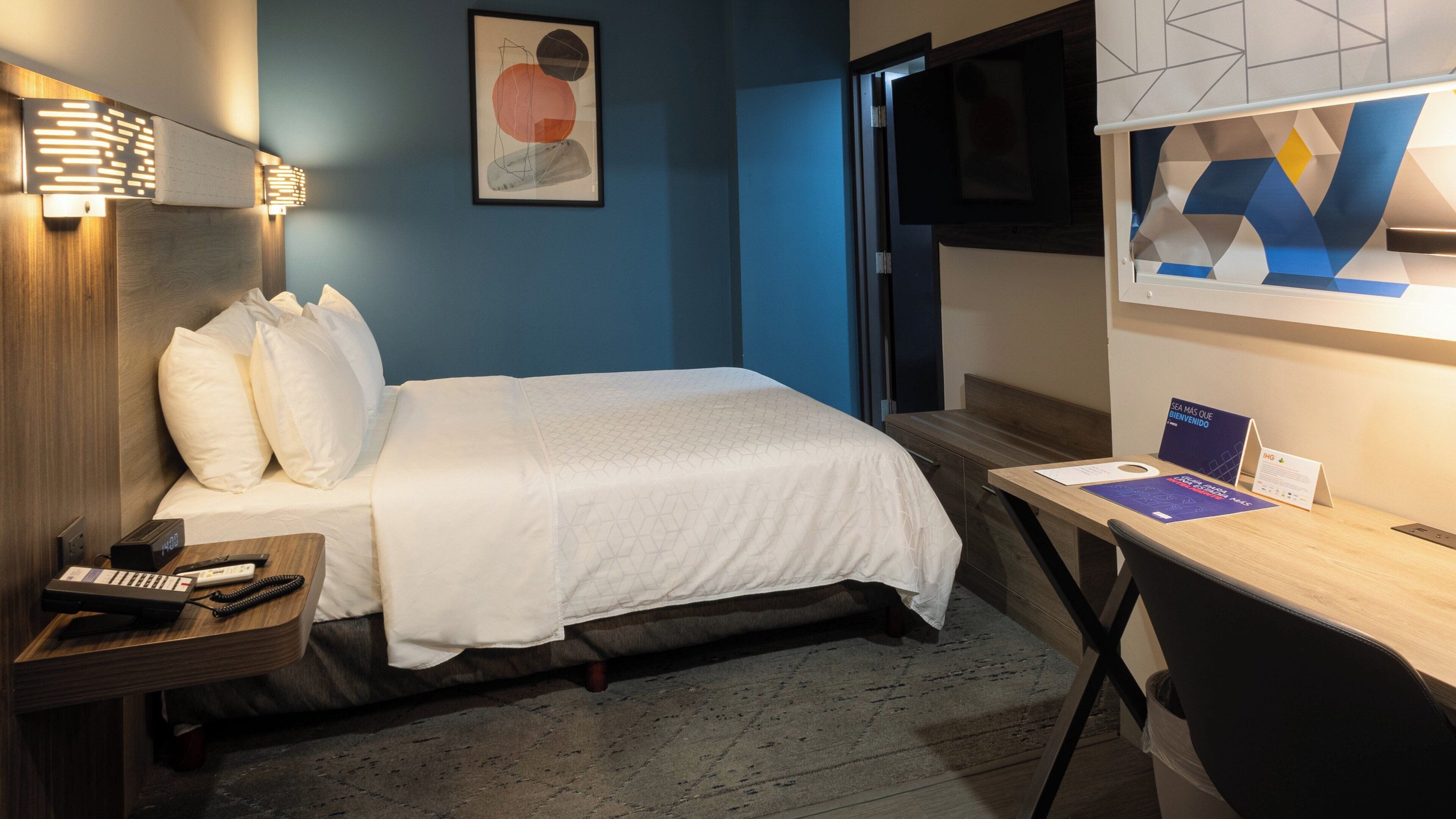 A hotel room with a bed, bedside table, desk, chair, and a TV on the wall.