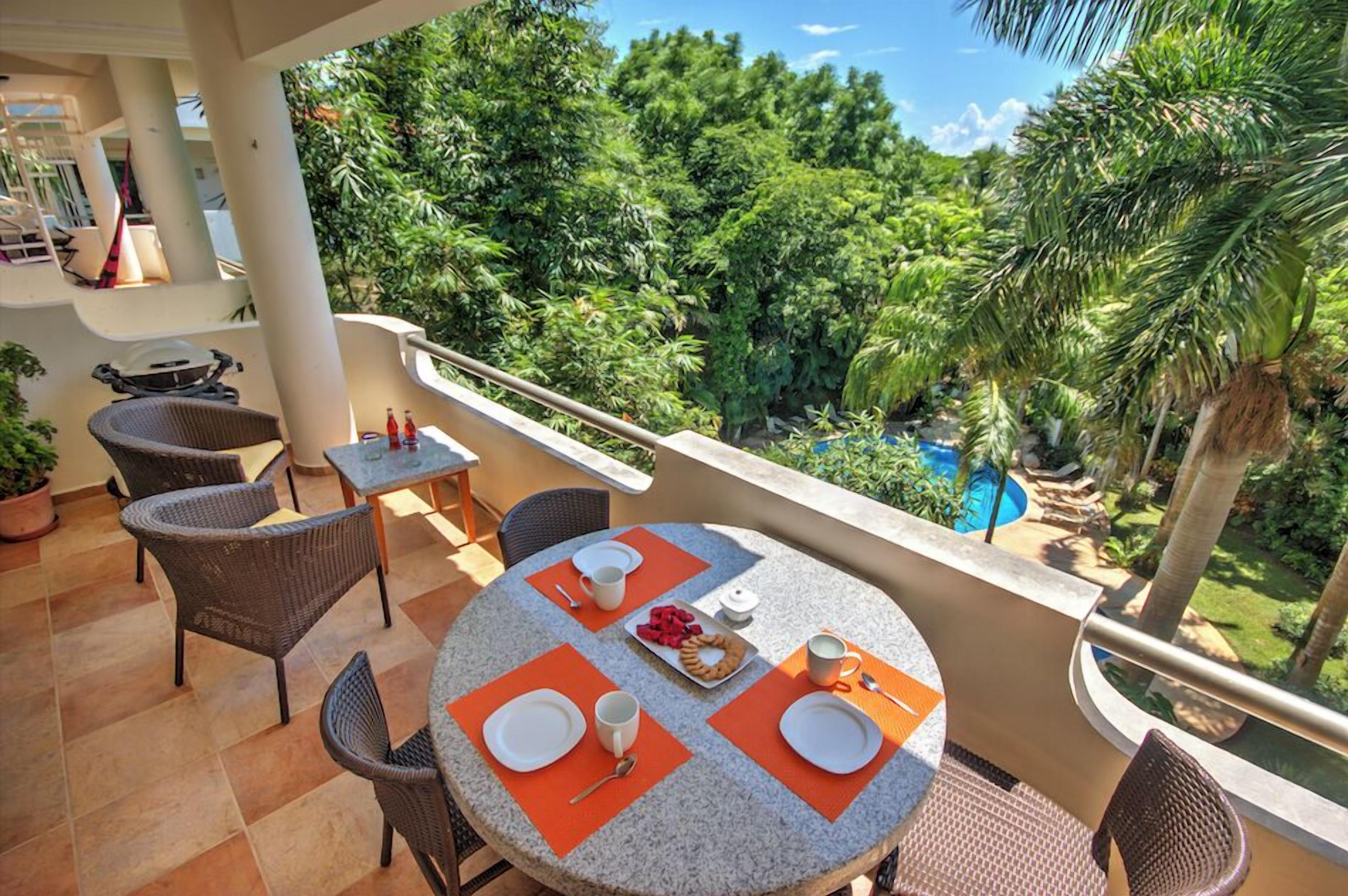 A balcony with a round table set for breakfast, surrounded by wicker chairs, overlooking a lush garden and a swimming pool.