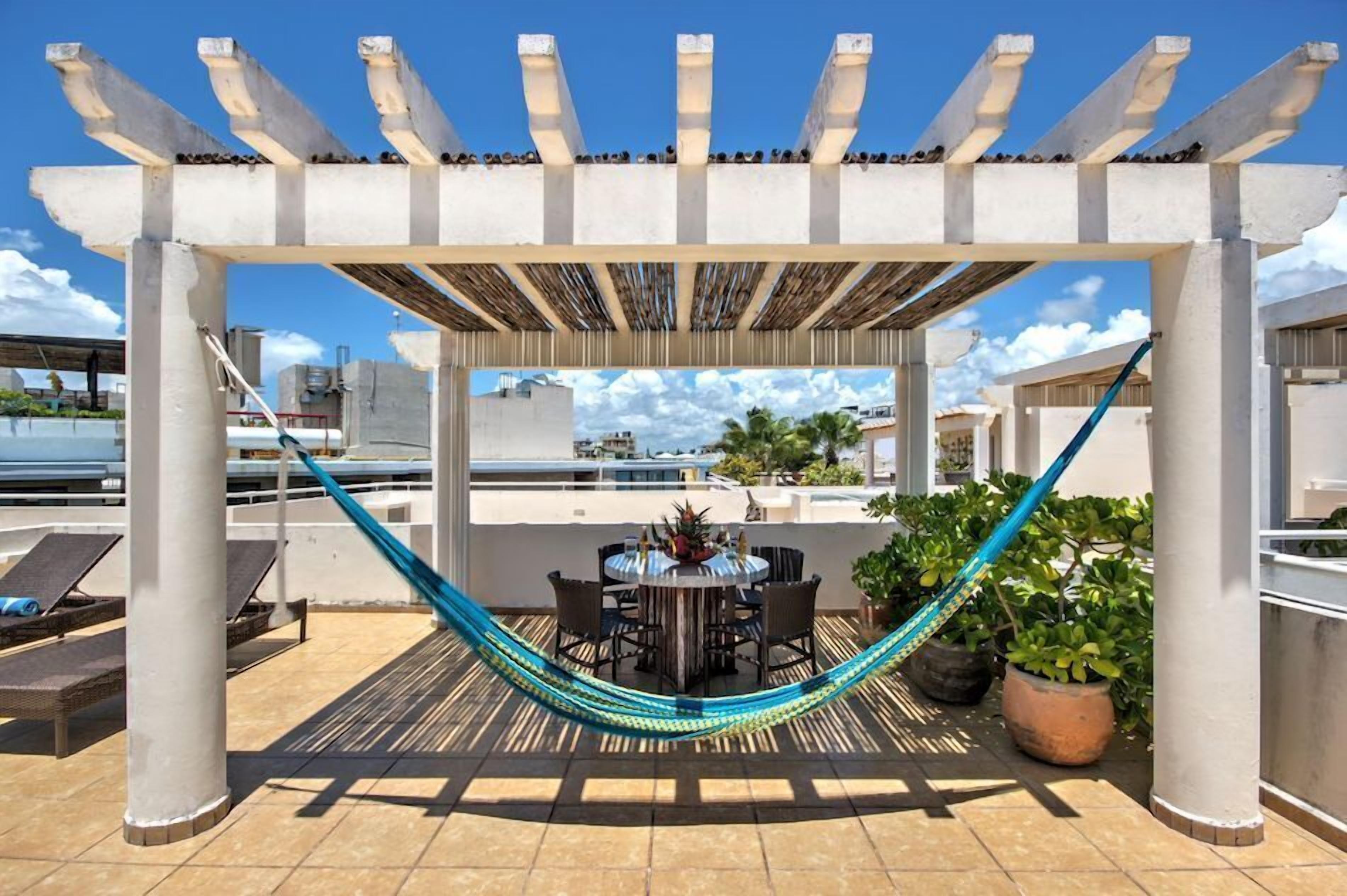 A rooftop patio with a pergola, a hammock, a dining table with chairs, and potted plants.