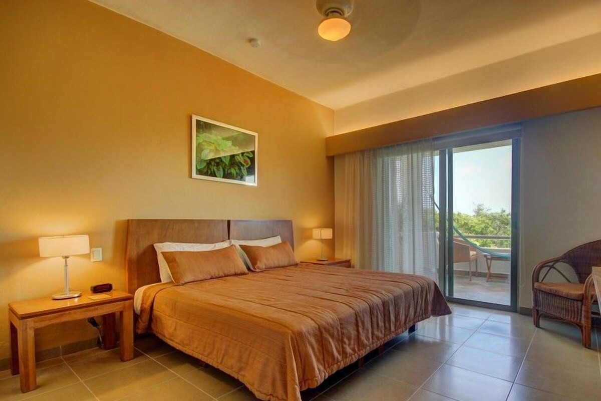 A hotel room with a large bed, two bedside tables, a chair, and a view of the outdoors through a sliding glass door.