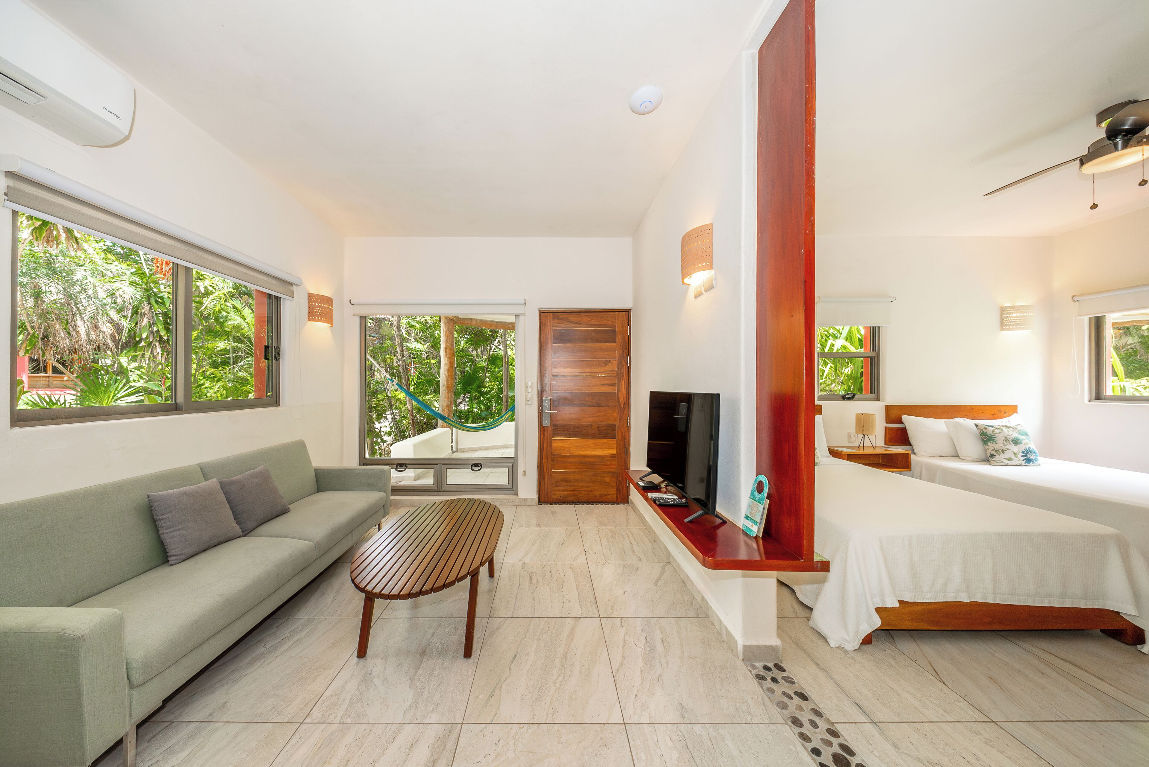 A modern room with a sofa, a wooden table, a bed, a TV, and a large window with a view of greenery.