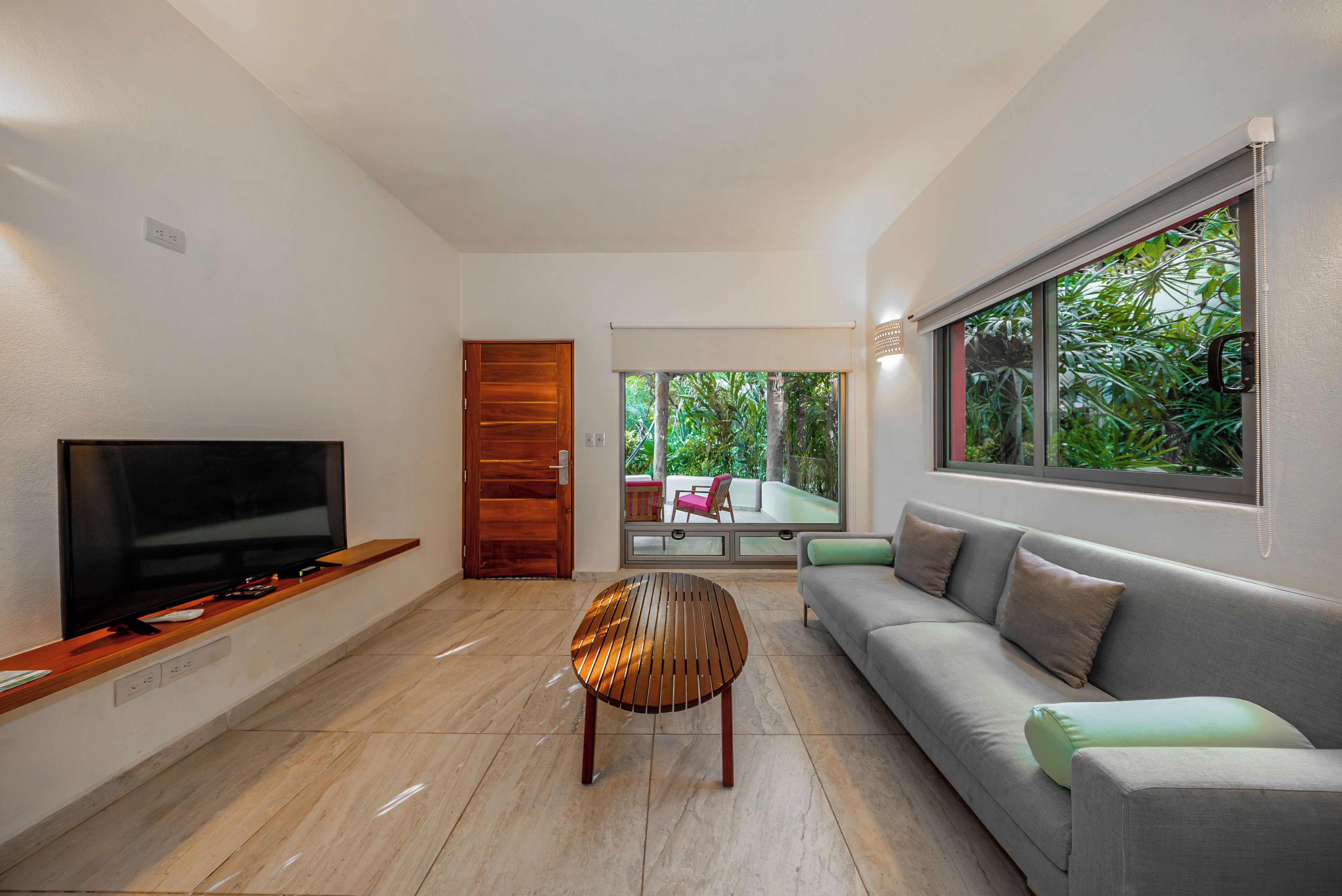 A modern living room with a flat-screen TV, a wooden coffee table, a grey sofa, and a large window with a view of greenery.