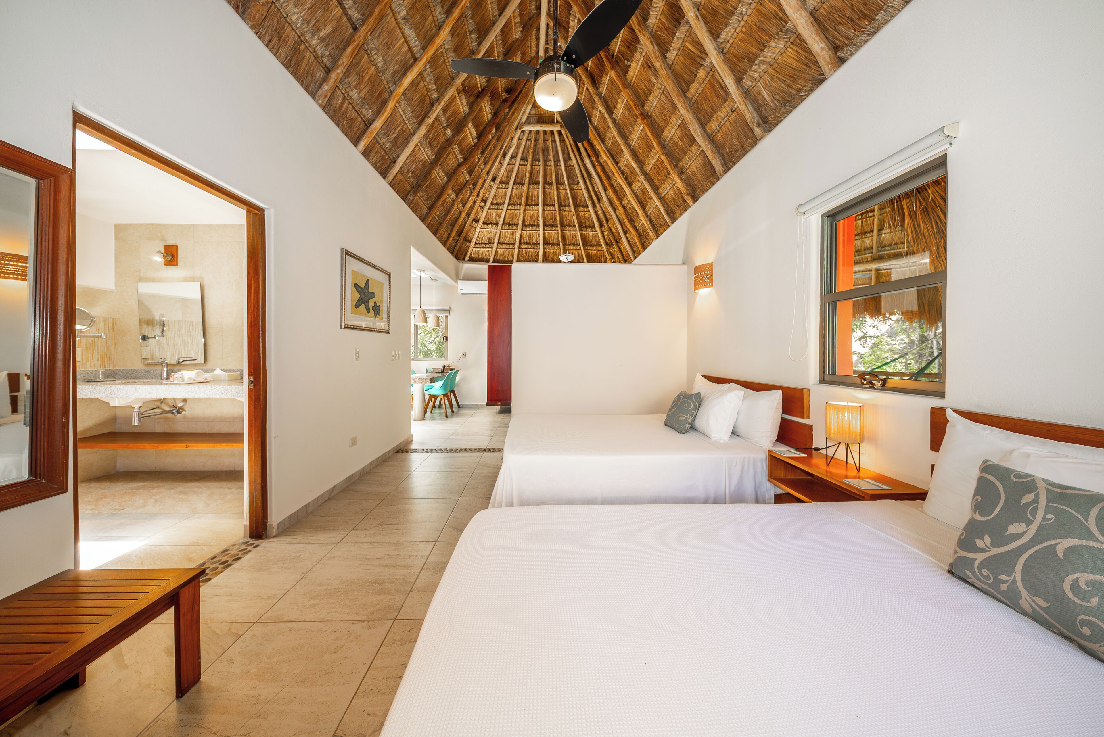 A hotel room with a thatched ceiling, two beds, a wooden bench, a bathroom with a mirror, and a window with a view.