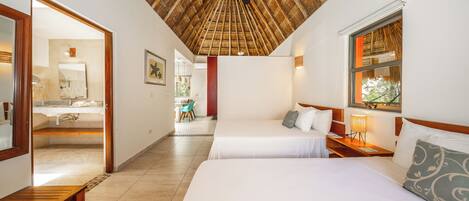 A hotel room with a thatched ceiling, two beds, a wooden bench, a bathroom with a mirror, and a window with a view.