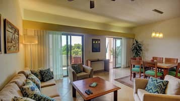 A spacious living area with a sofa, coffee table, and dining table. There is a ceiling fan, a lamp, and a plant.