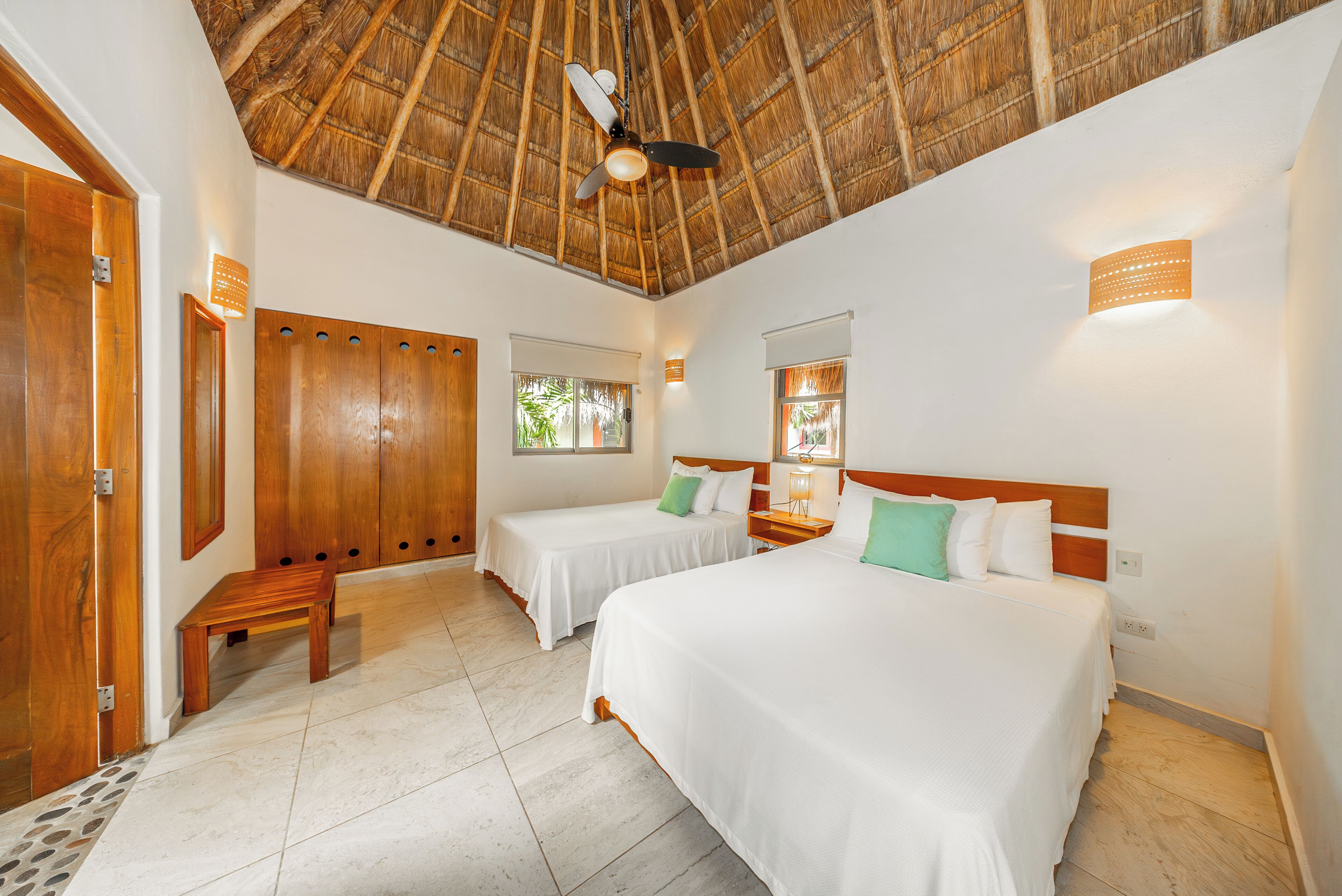 A room with two beds, a wooden bench, a ceiling fan, and a thatched roof.