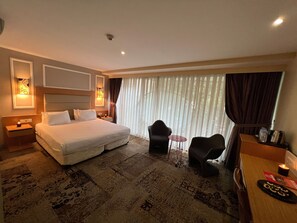 Ein Hotelzimmer mit einem großen Bett, Nachttischen, einem Schreibtisch mit Stuhl, einem kleinen Tisch mit Stuhl und einem großen Fenster mit Vorhängen.