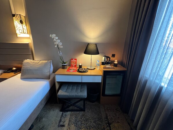 Une chambre d’hôtel avec un lit, un bureau doté d’une lampe, une petite table avec une plante et un livre, et une table de chevet avec un téléphone et une bougie.