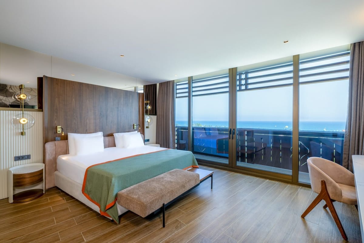 Una habitación de hotel moderna con una cama grande, un banco, una silla y un balcón con vista al mar.