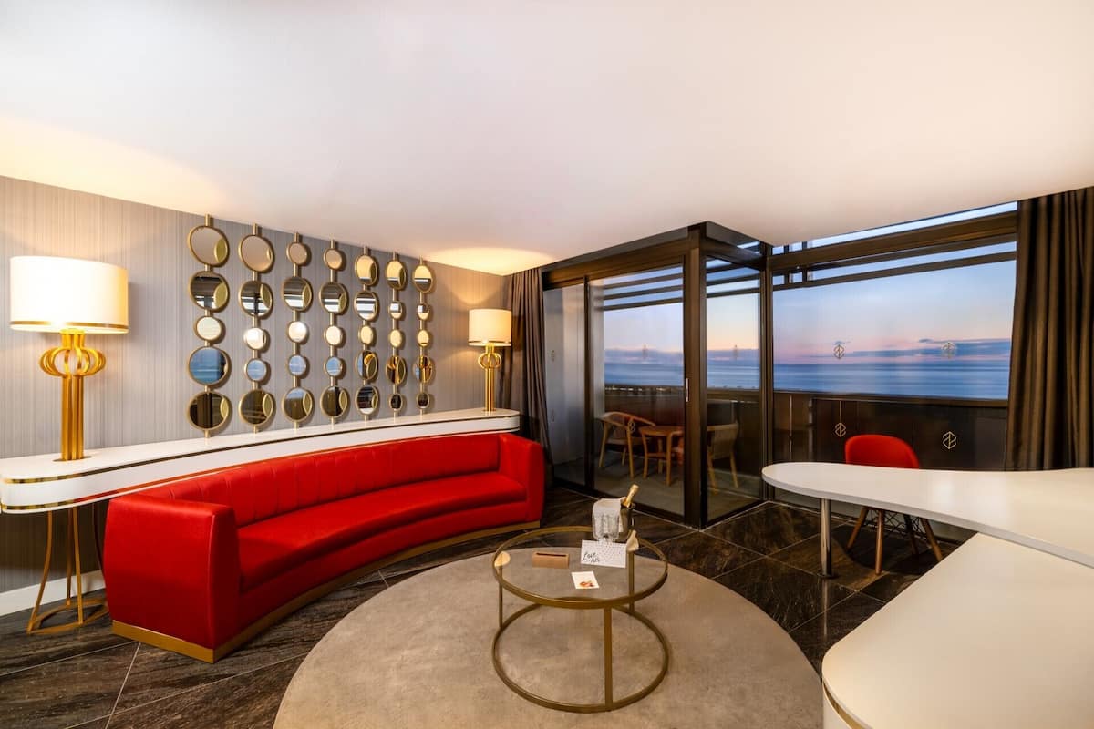 Una habitación de hotel moderna con un sofá rojo, una mesa de centro redonda de vidrio, un escritorio blanco y una pared de espejos circulares. Hay un balcón con vistas al océano.