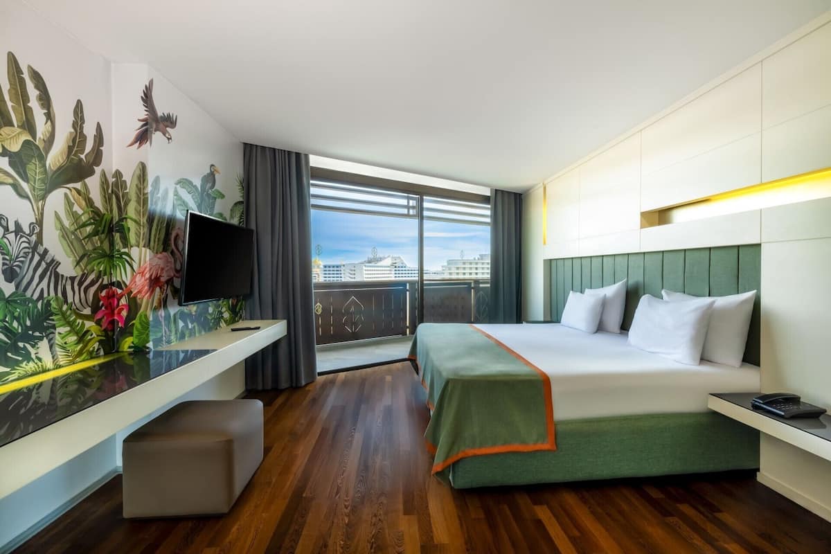 Habitación de hotel con cama, escritorio con televisor, balcón y vista a la ciudad, además de un mural grande en la pared.