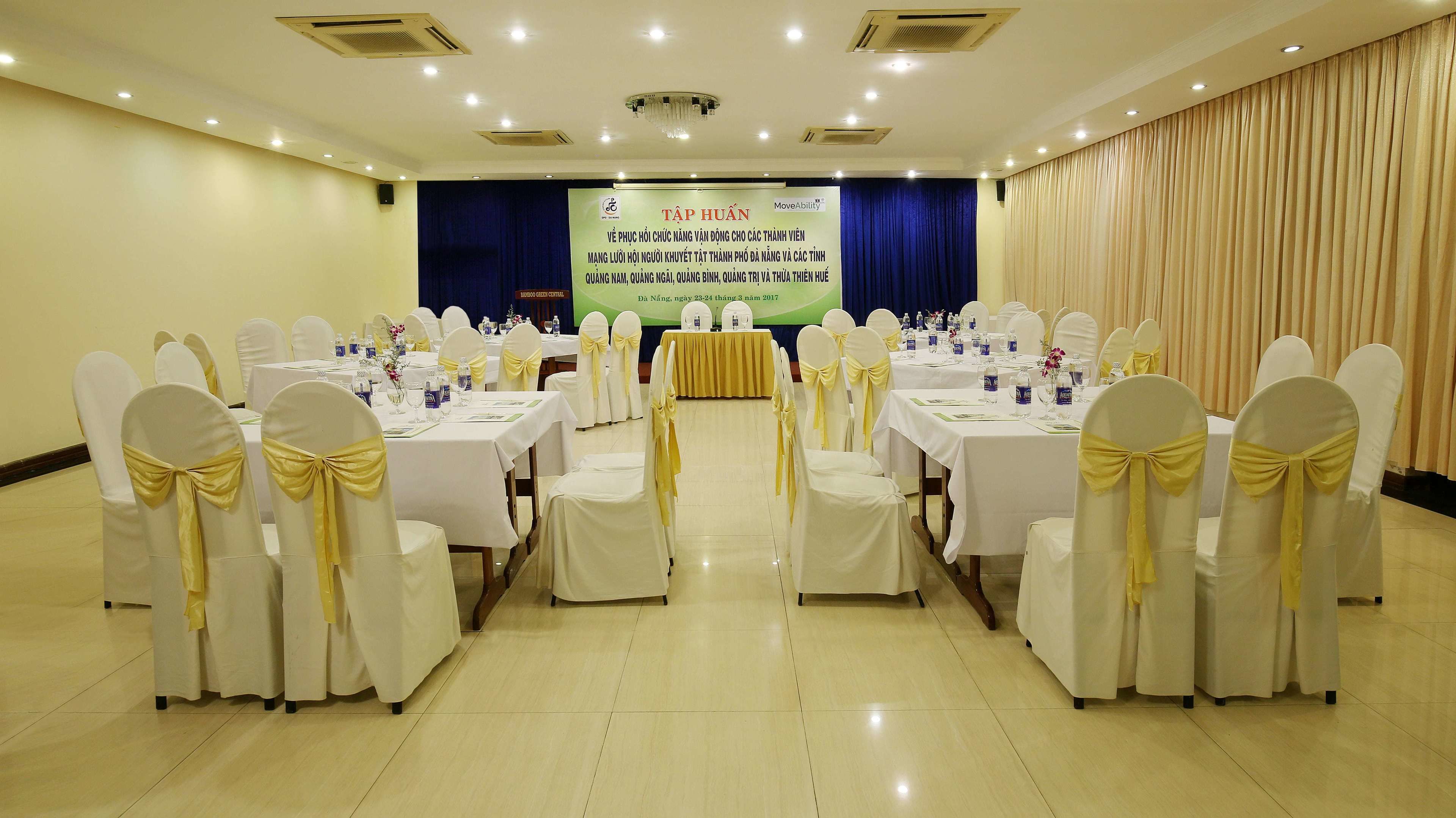 A conference room with tables set for a meeting, white chairs with yellow sashes, a stage with a banner, and a curtain backdrop.
