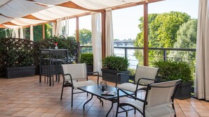 A covered outdoor seating area with a table and chairs, potted plants, and a view of a bridge and trees.