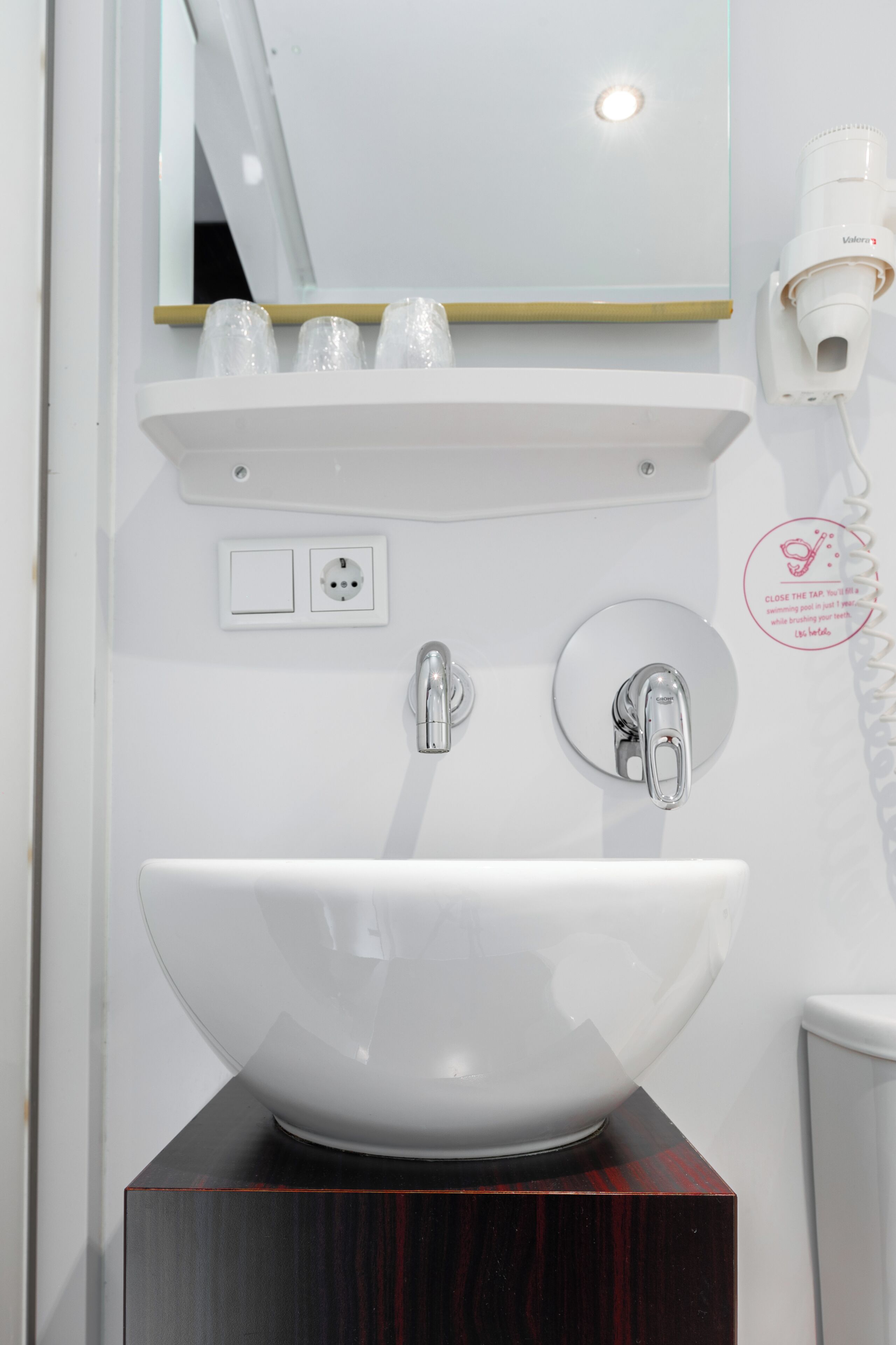 Een witte wasbak met een groot bassin, een spiegel erboven en een klein plankje met bekers.