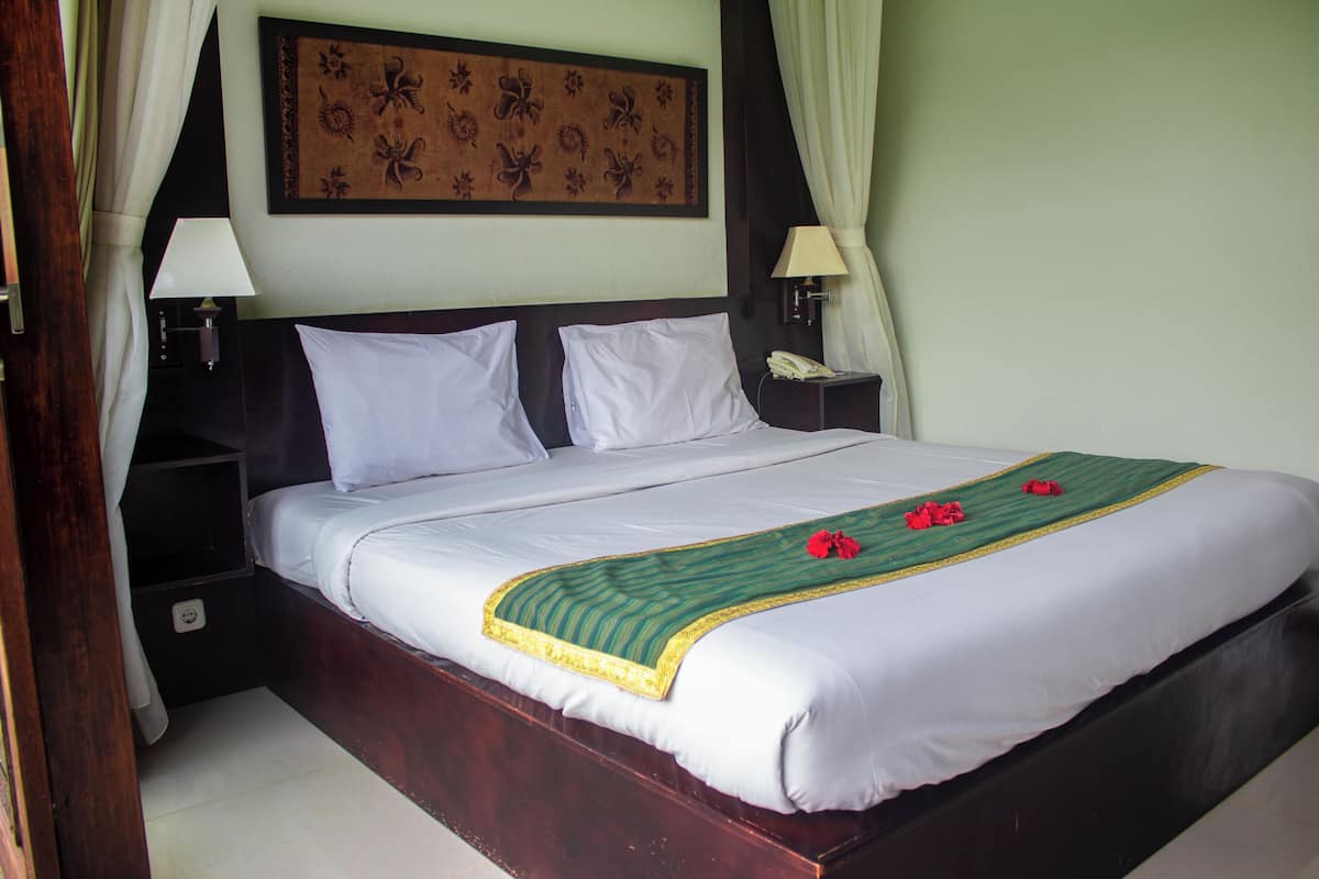 Una cama bien hecha con sábanas blancas, una colcha verde y dorada, y flores rojas. Hay dos mesitas de noche con lámparas, un teléfono en la pared y un adorno decorativo sobre la cama.