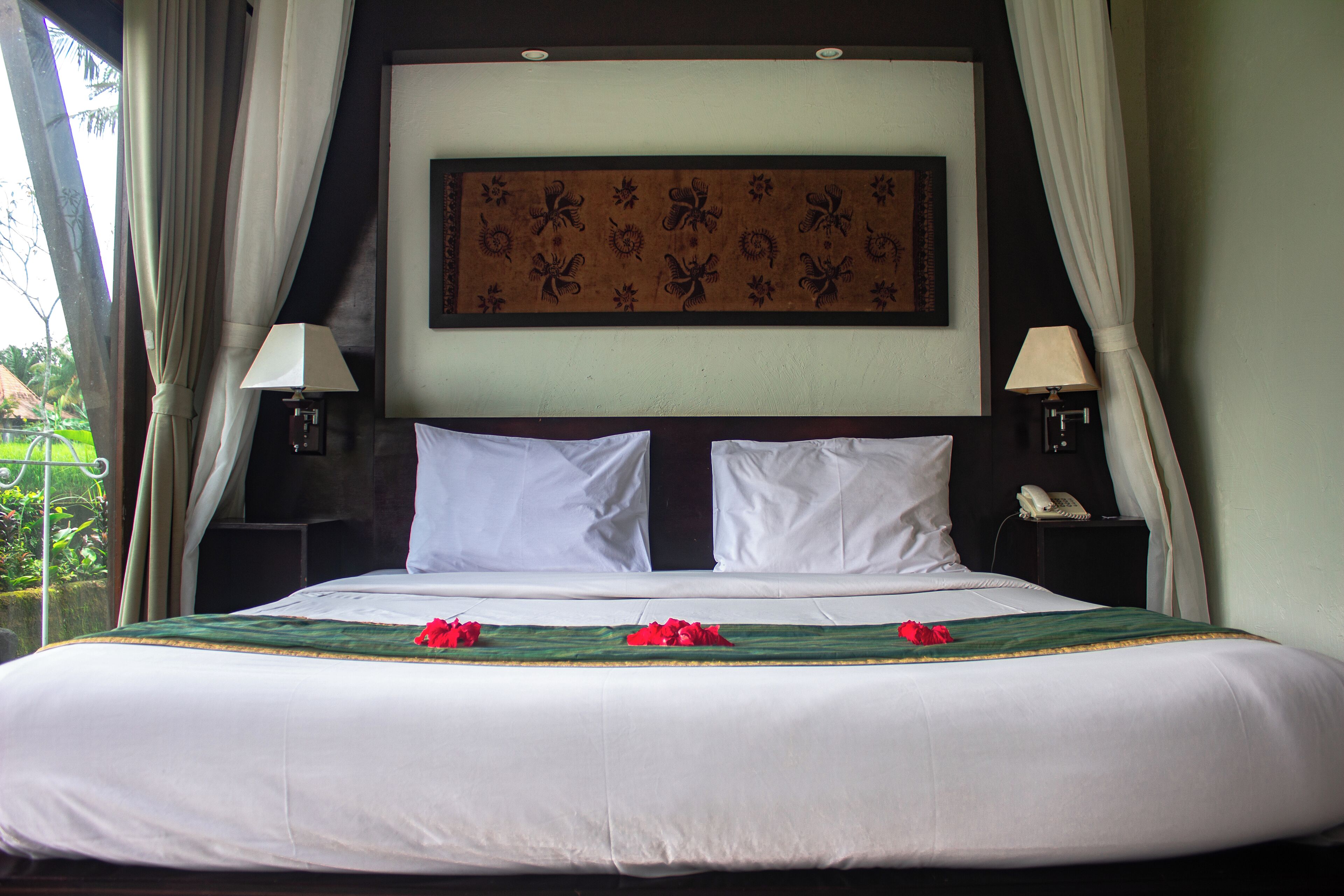 A neatly made bed with white linens and a green bedspread, two white pillows, and red flowers on the bed. There are two bedside tables with lamps and a telephone. A large floral artwork is on the wall above the bed.