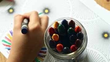 Uma mão segurando uma caneta, desenhando em um papel com padrão. Um pote cheio de marcadores coloridos está em primeiro plano.