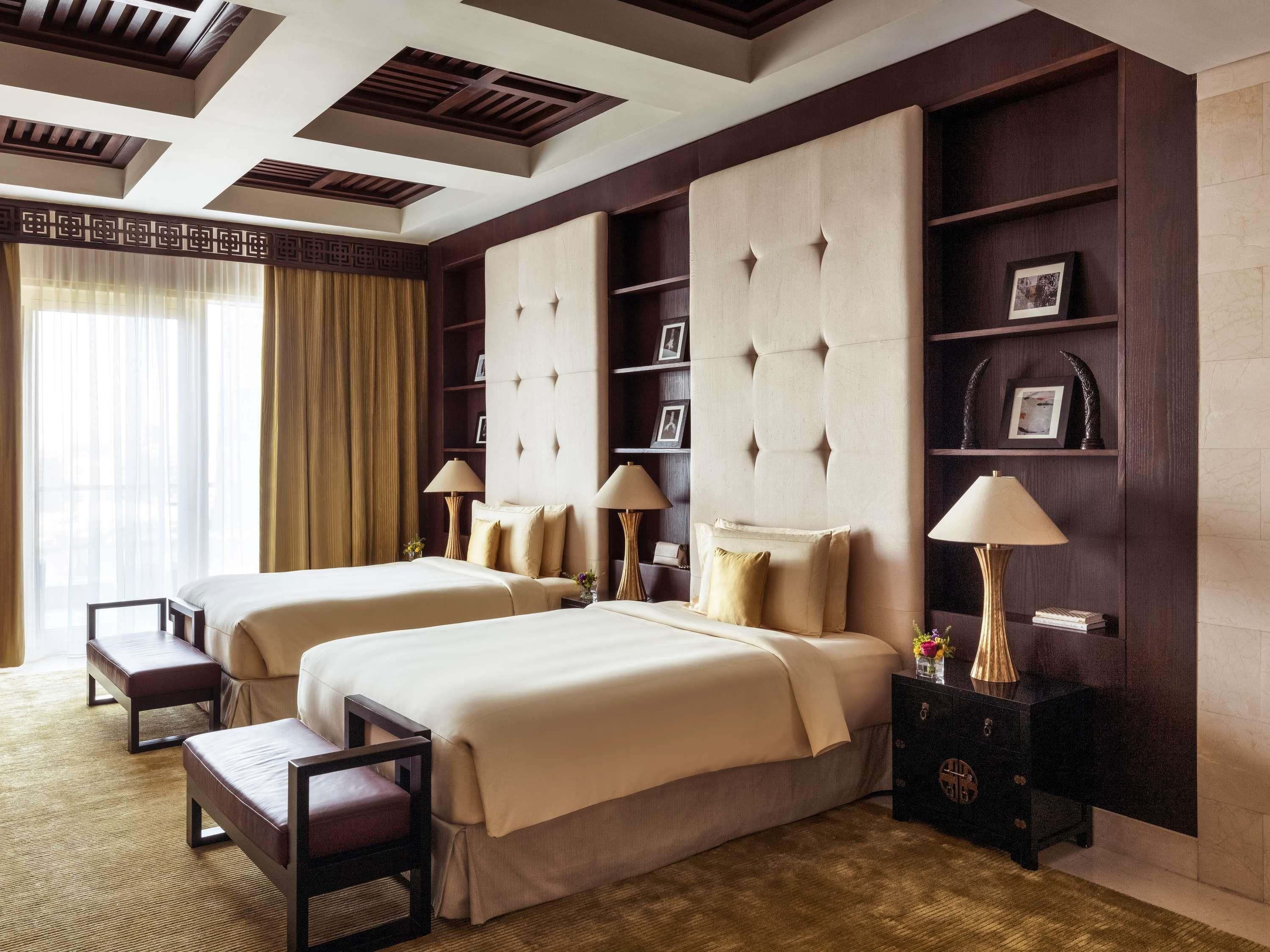 A hotel room with two beds, a large window with curtains, a bookshelf, and a decorative wall panel.