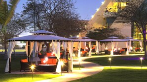 An outdoor seating area with tables and chairs under canopies, a pathway, and a building in the background.