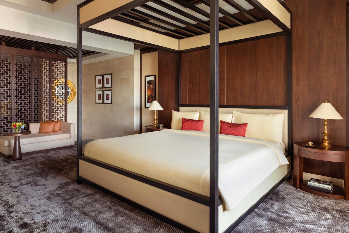 A four-poster bed with a canopy, a sofa, a side table with a lamp, and a decorative wall panel.