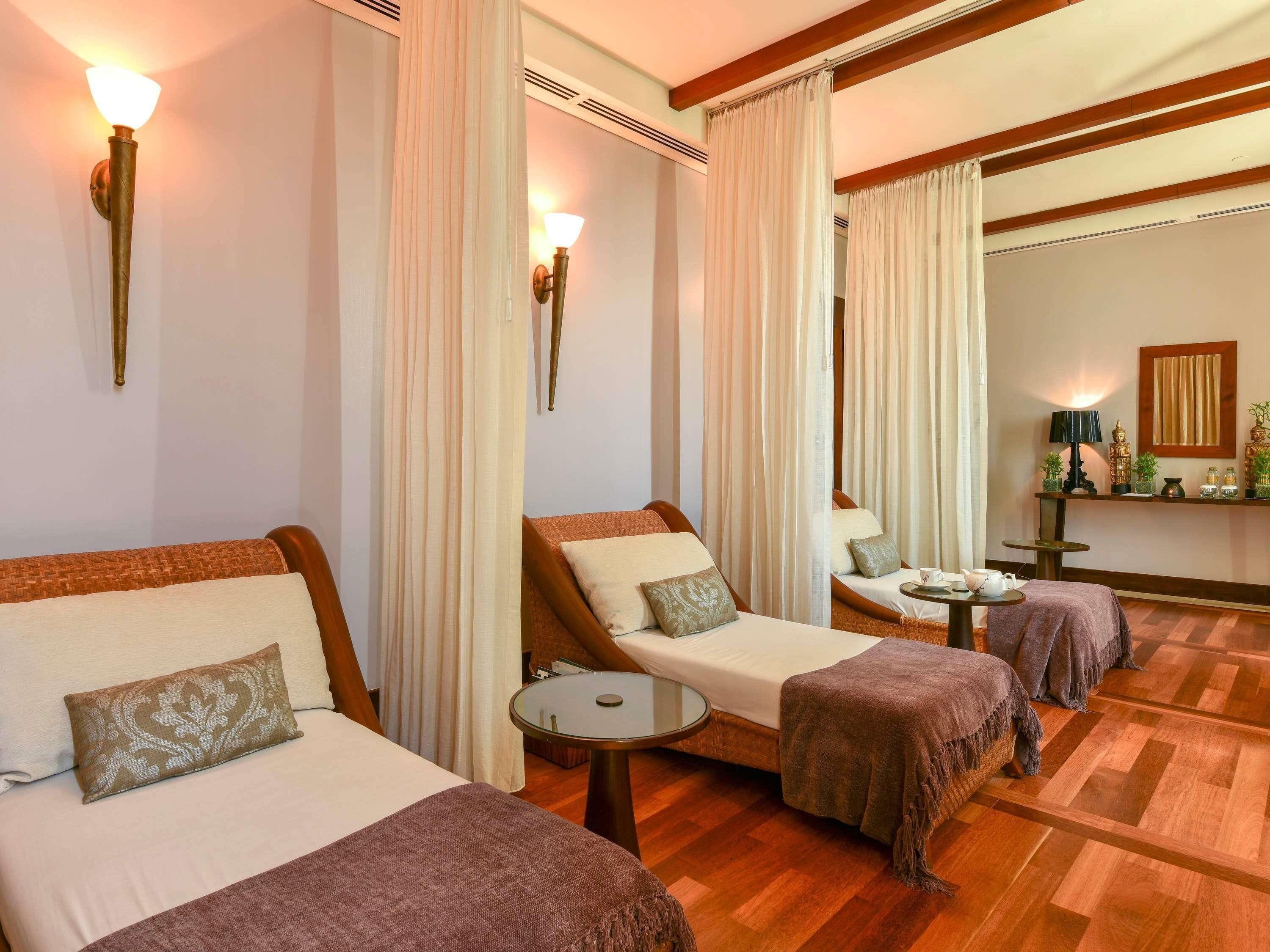 A hotel room with two beds, a small table, a mirror, and a decorative shelf with plants.