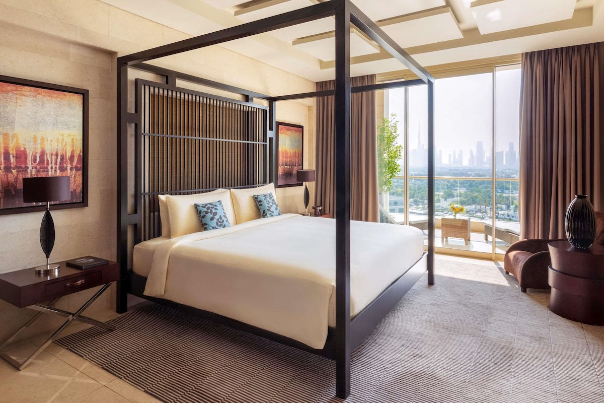 A modern hotel room with a four-poster bed, a city view, and a large painting on the wall.