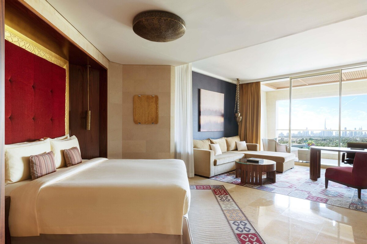 A spacious hotel room with a large bed, a sofa, a coffee table, and a view of the city skyline.