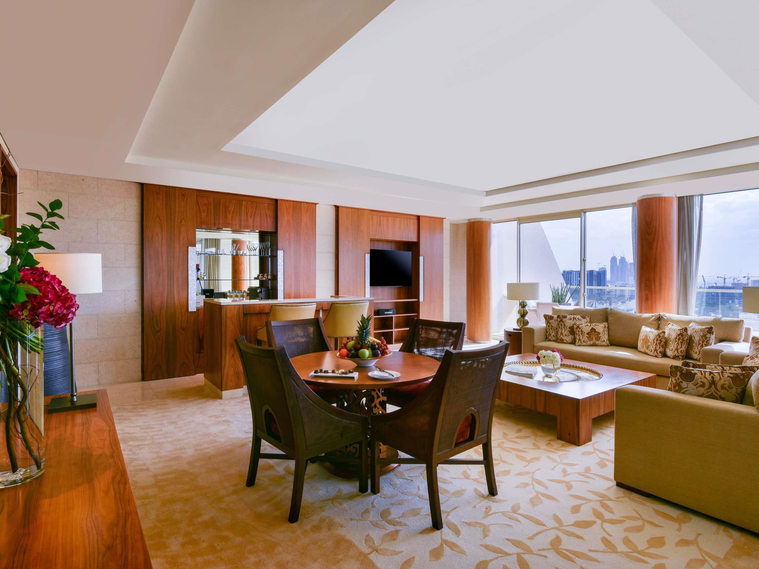 A modern hotel room with a dining area, a sofa set, and a large window offering a city view.