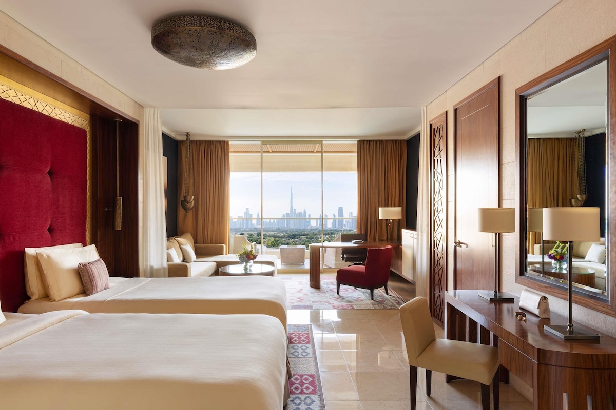 A hotel room with two beds, a desk, a chair, a large mirror, and a view of a city skyline.