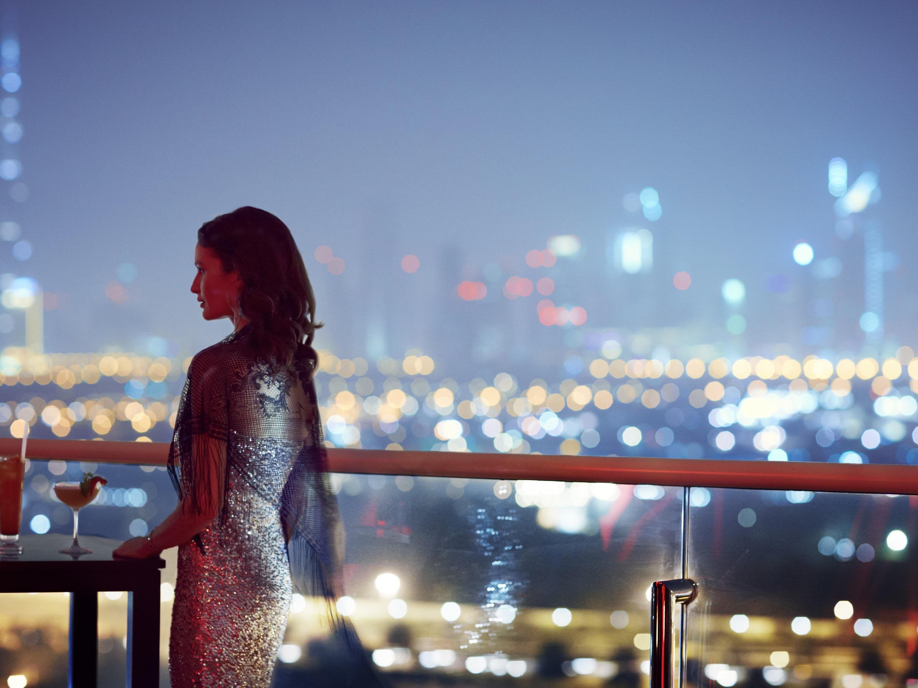 A person in a shiny dress standing by a railing with a cityscape view in the background.