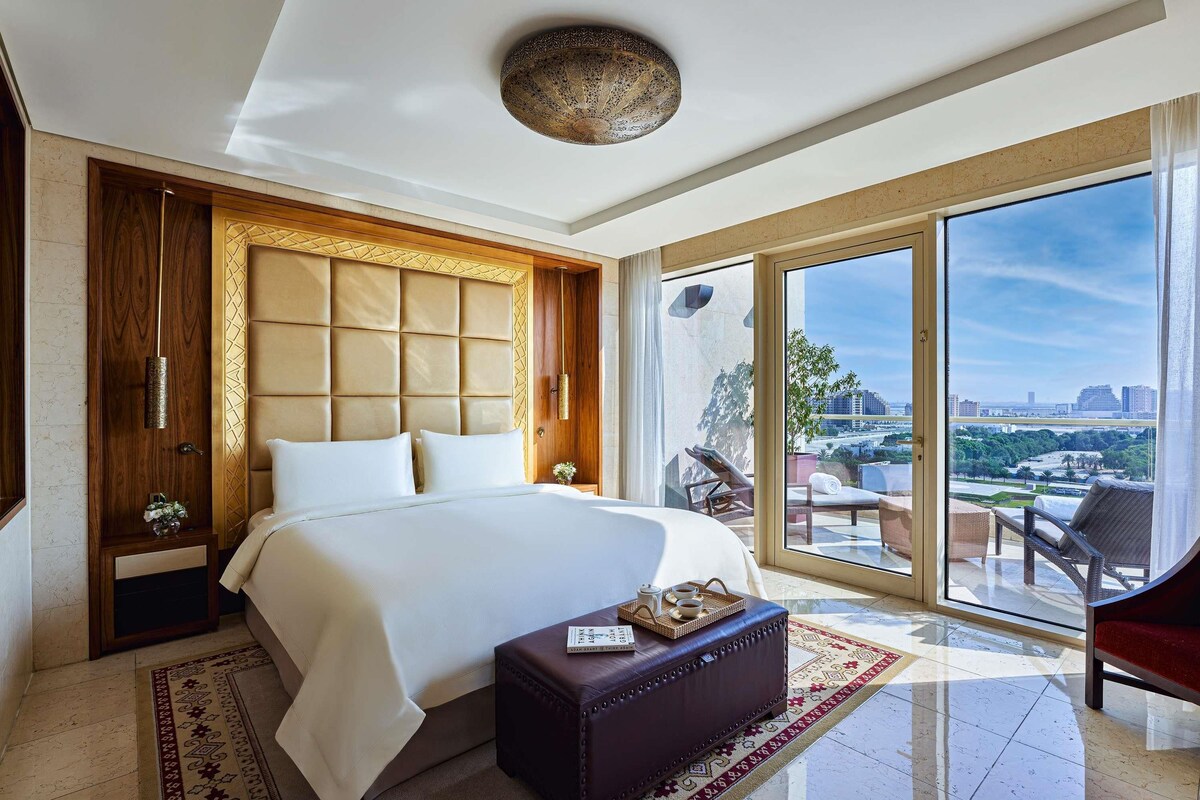 A modern hotel room with a large bed, a wooden headboard, a view of the city, and a balcony with outdoor furniture.