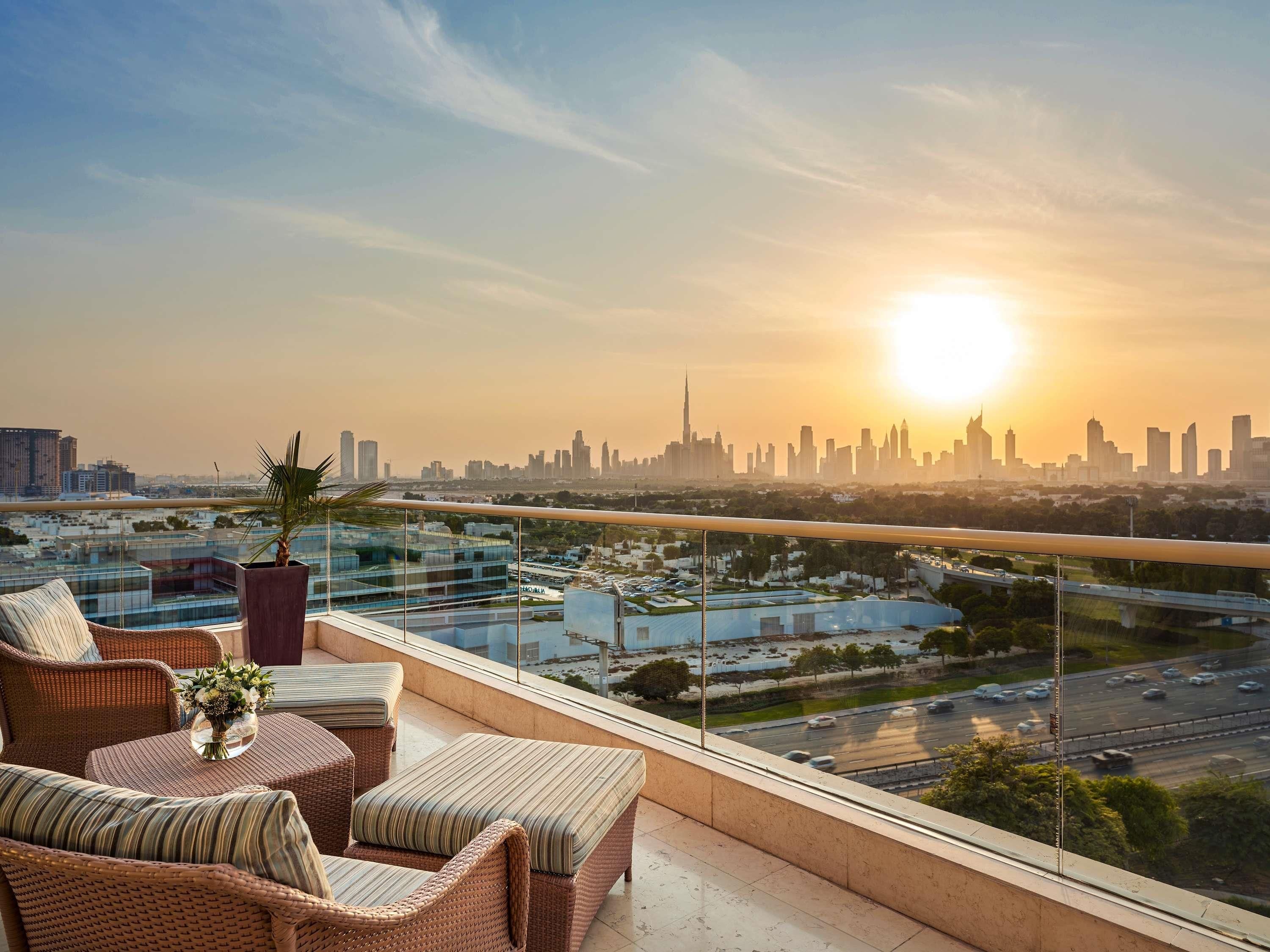 A balcony with wicker furniture, a potted plant, and a view of a city skyline at sunset.
