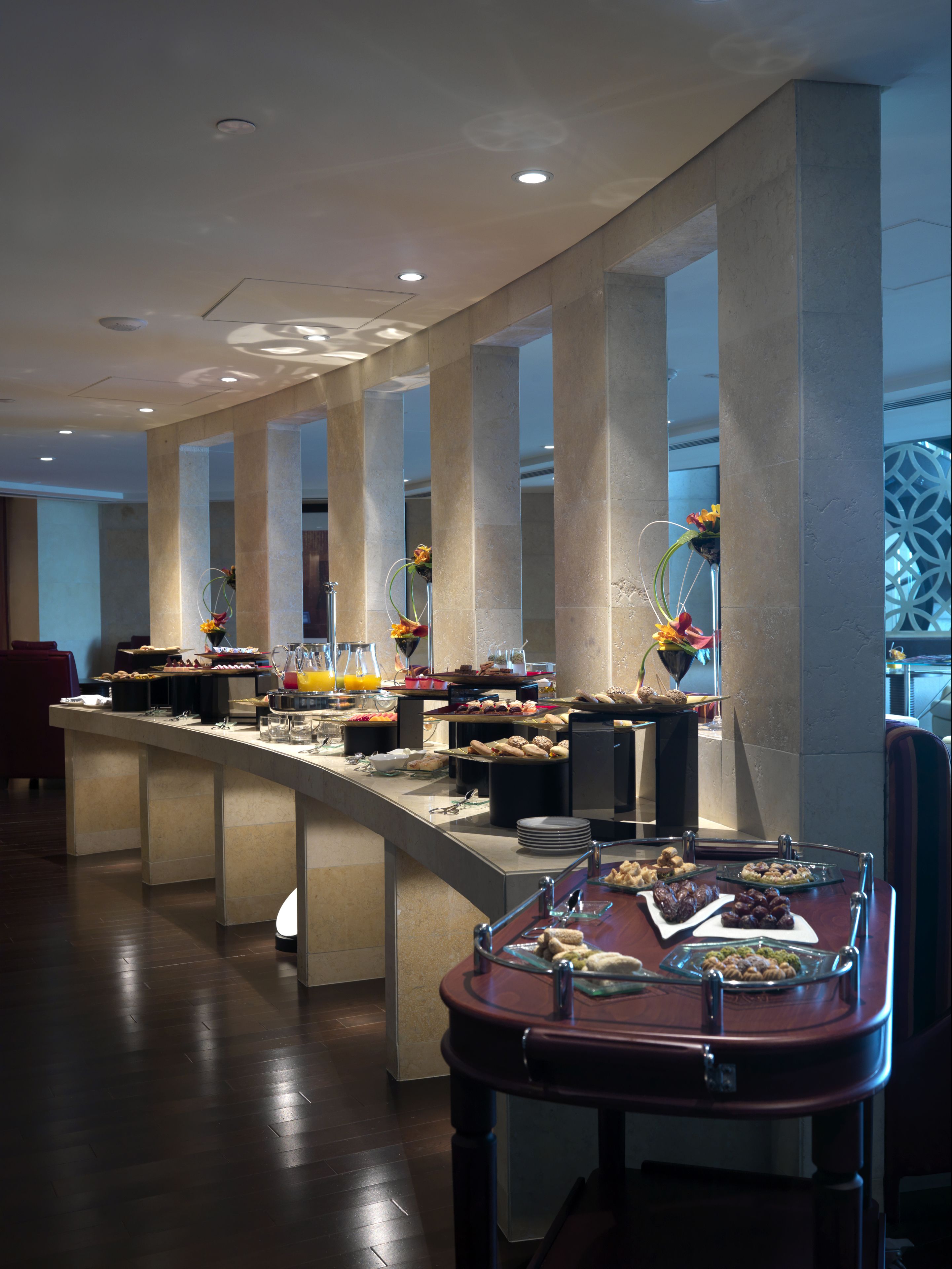 A buffet setup with various food items and drinks, a modern interior with large columns, and a patterned wall in the background.