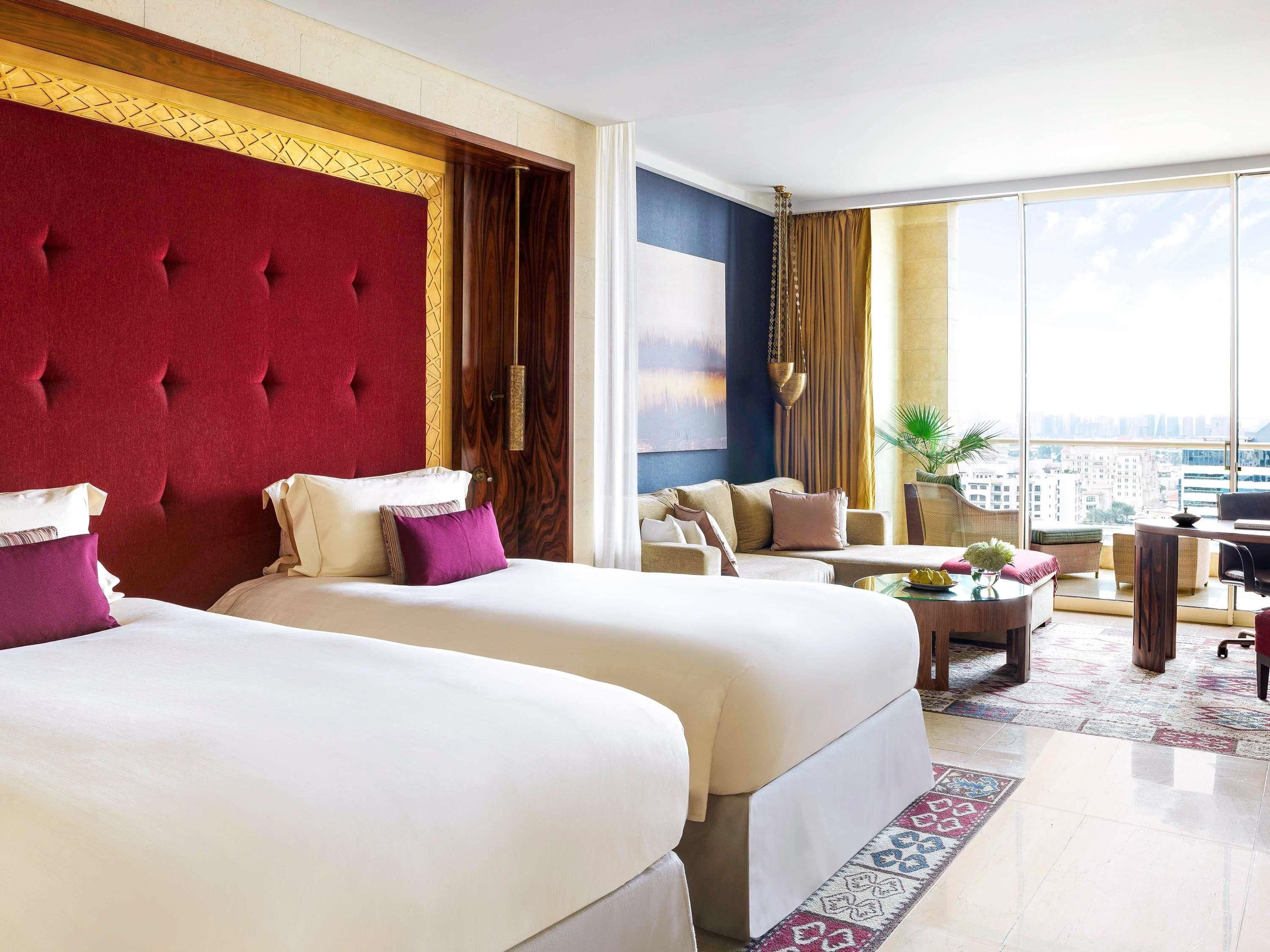 A hotel room with two beds, a sofa, a dining table, and a large window offering a city view.