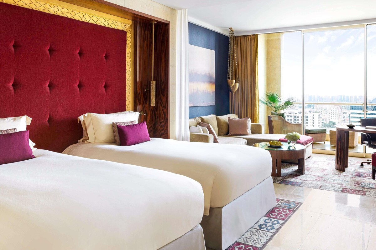 A hotel room with two beds, a sofa, a dining table, and a large window offering a city view.