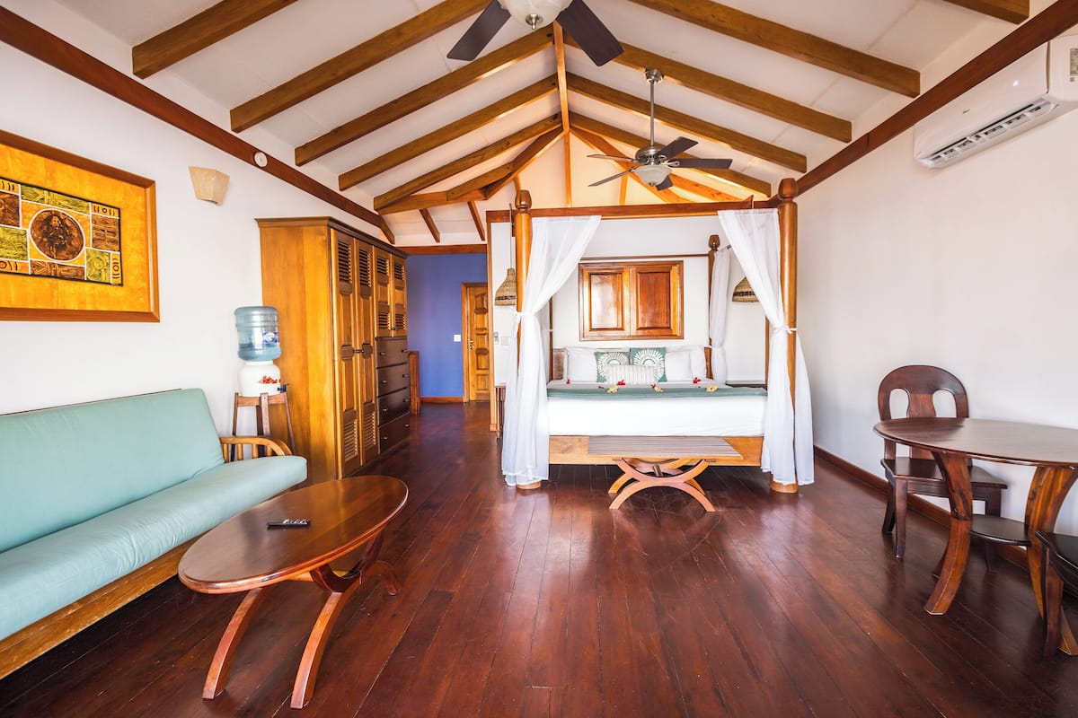 A room with a wooden ceiling, a bed with a canopy, a sofa, a table, and a chair.