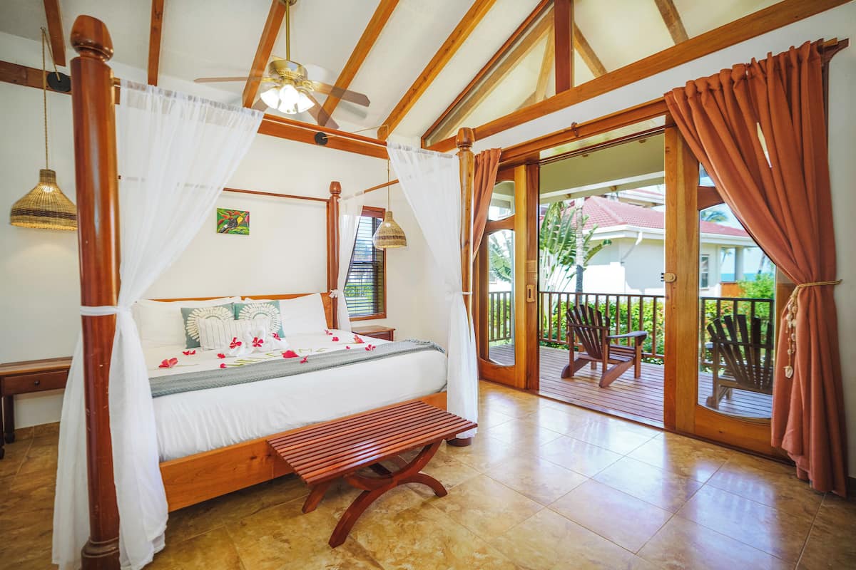 A spacious bedroom with a large bed, wooden ceiling beams, a ceiling fan, and a balcony with wooden chairs.