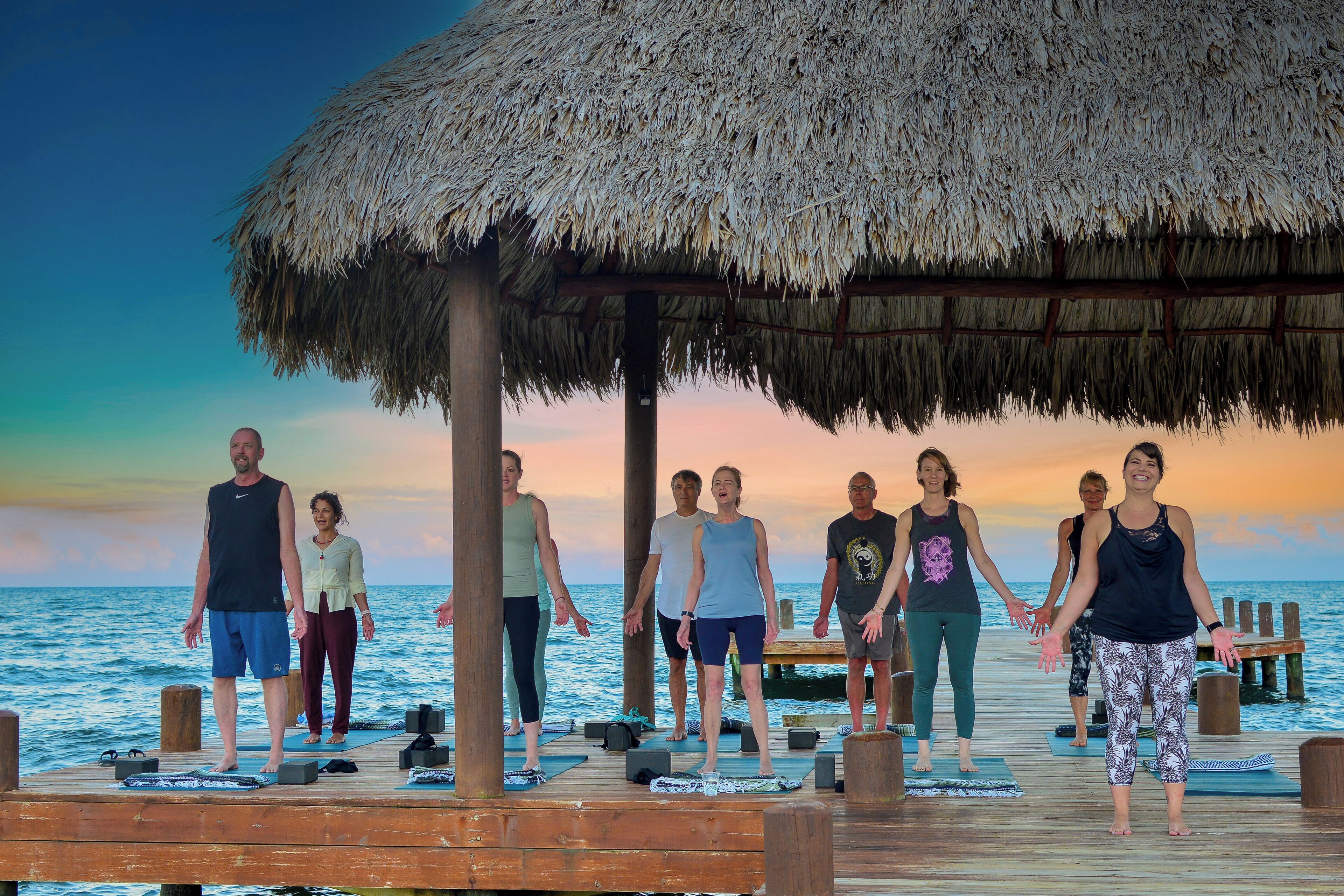 A group of people performing a yoga exercise on a wooden deck with a thatched roof, overlooking the sea.