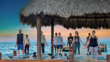 A group of people performing a yoga exercise on a wooden deck with a thatched roof, overlooking the sea.