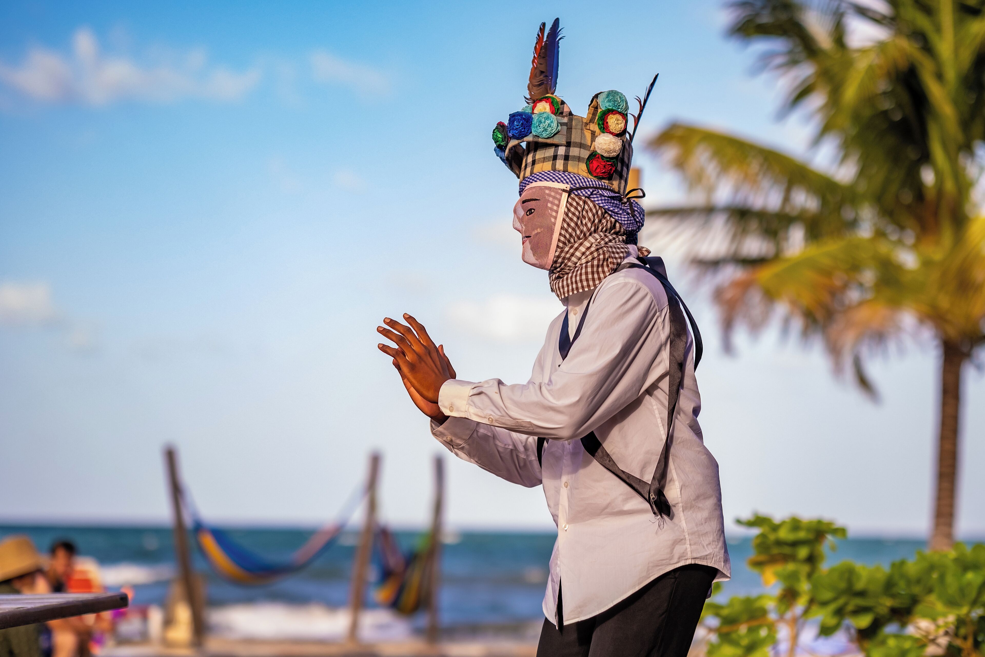 Una persona con camisa blanca y pantalones negros de pie en una playa con palmeras y el océano al fondo.