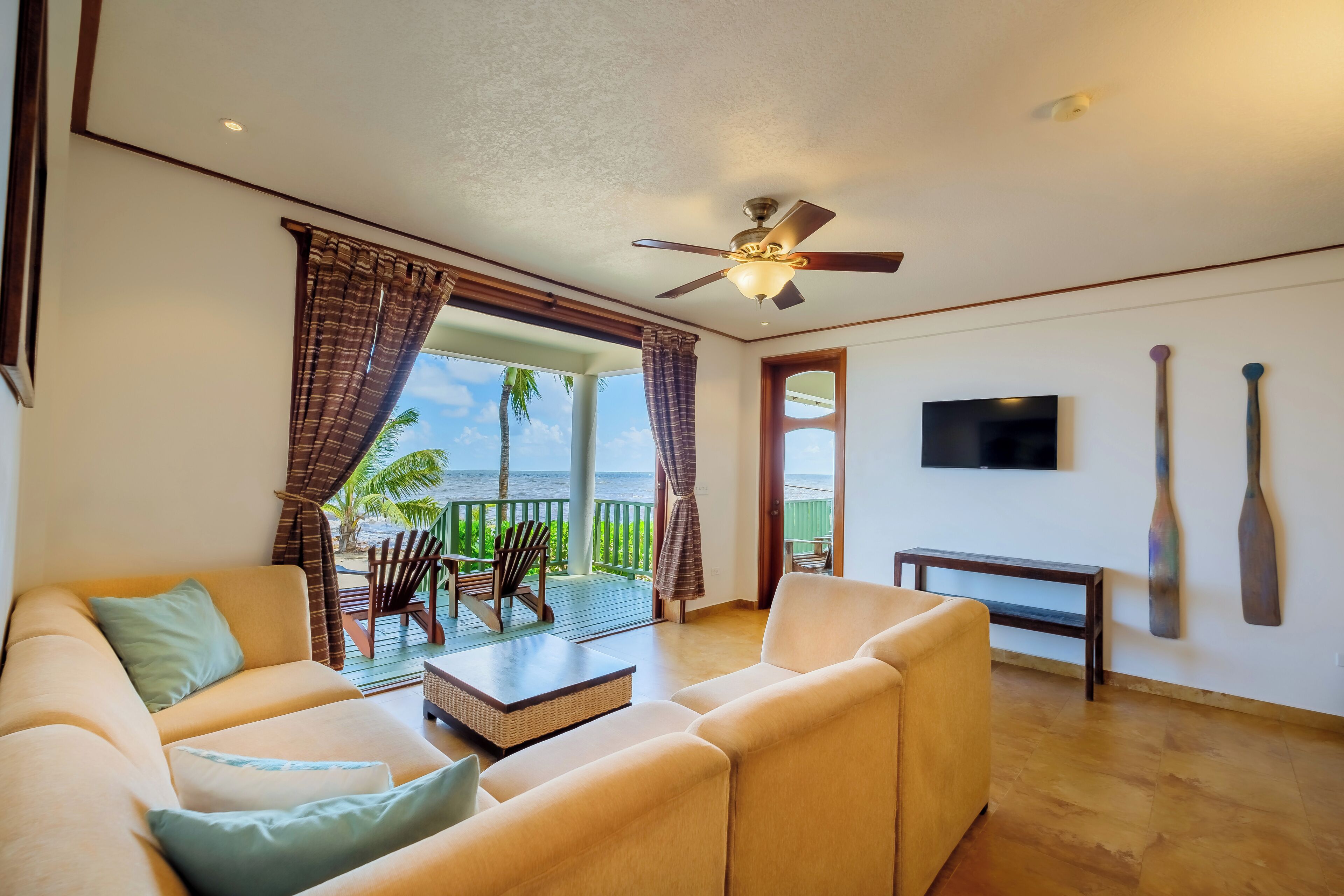 A spacious living room with a sofa, a coffee table, and a TV mounted on the wall. There is a balcony with a view of the ocean and palm trees.
