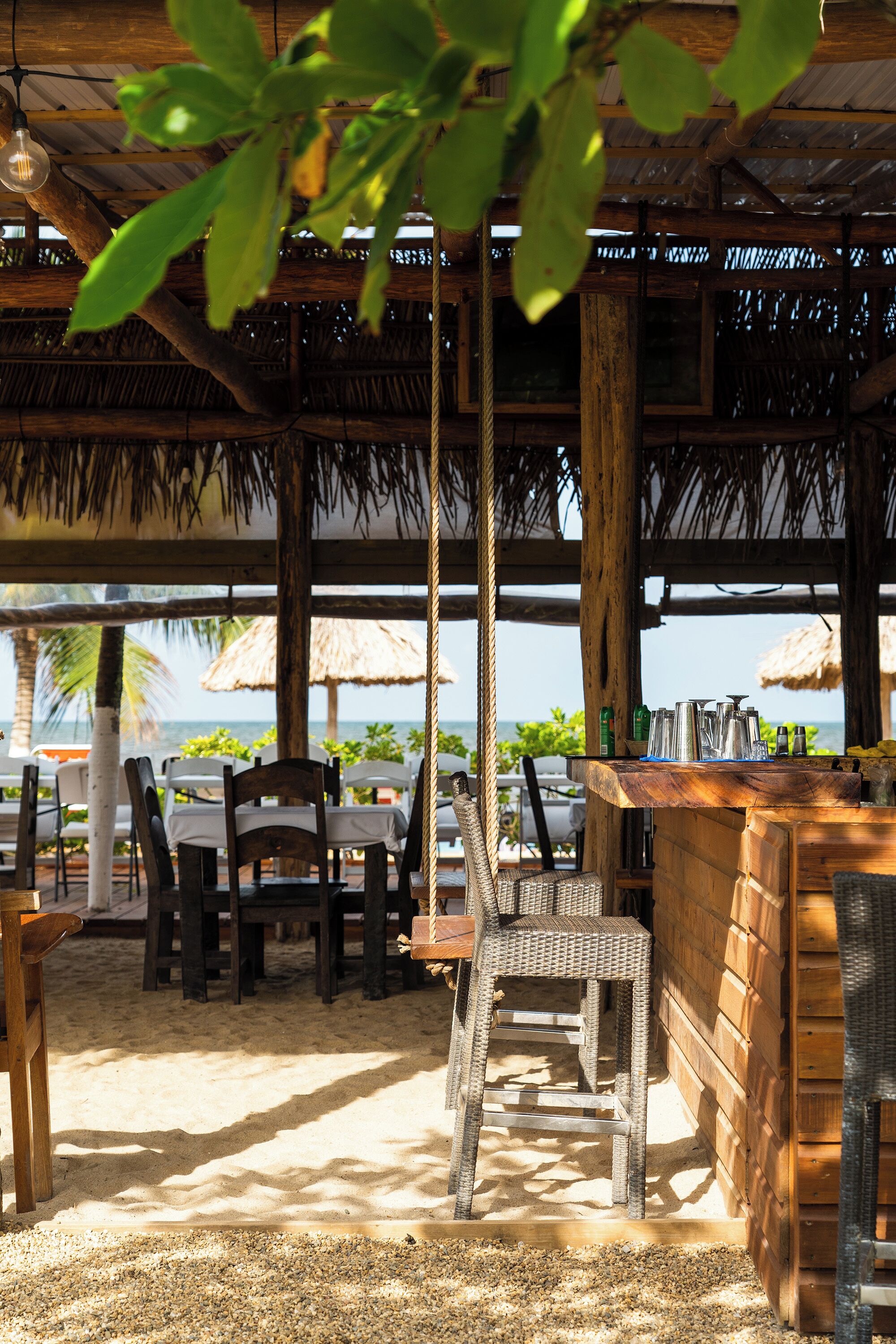 A beachside restaurant with wooden tables and chairs, a bar area, and thatched umbrellas.